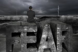 Businessman sitting on old concrete fear word, facing dark storm ocean.